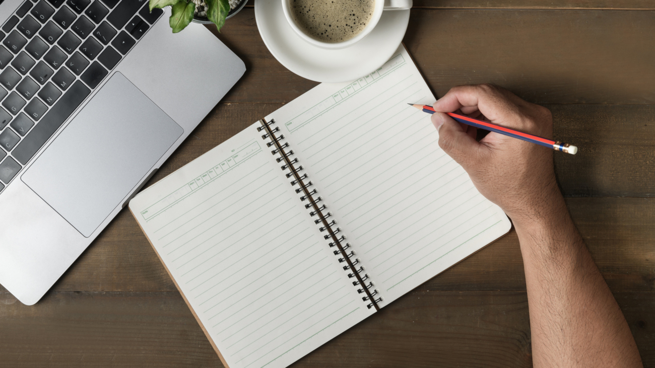 Businessman writing on blank notebook with hot coffee and laptop on wood table, top view